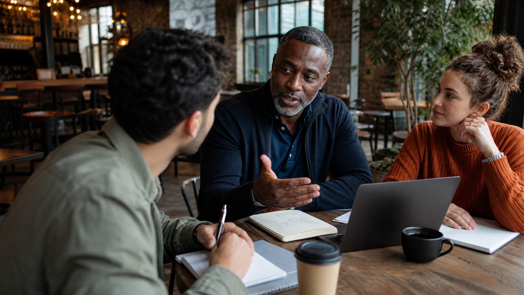 A business leader talking with two students over notebooks and a laptop at a coffee shop table.