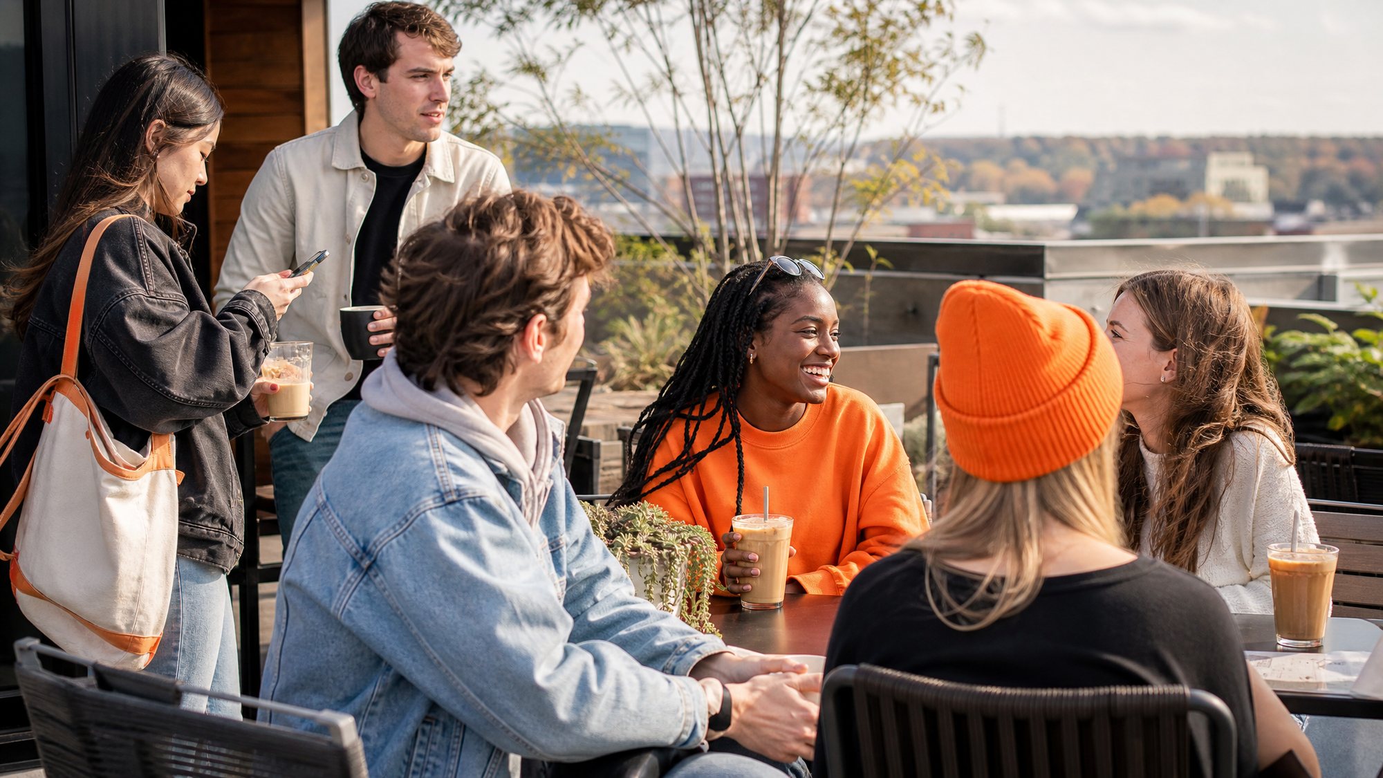 Students talking over coffee at tables on a rooftop terrace, with the city behind them.